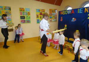 Pani dyrektor Maria Królikowska trzyma w ręku dużą żółtą kredkę, którą przykłada chłopcu do lewego ramienia.